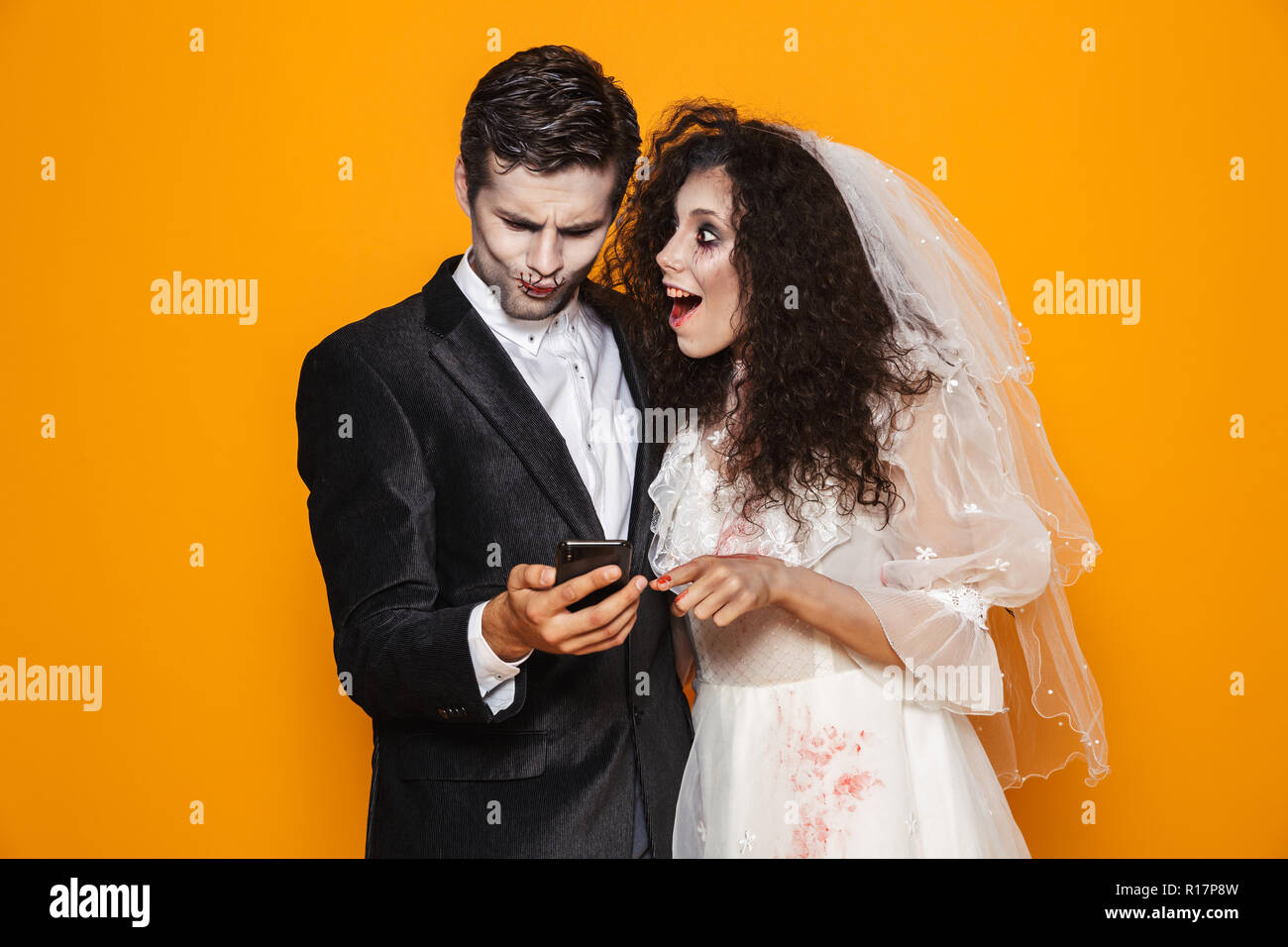Photo de beau couple marié mariée zombie et portant des vêtements de mariage et maquillage d'halloween à l'aide de smartphones fond jaune Banque D'Images