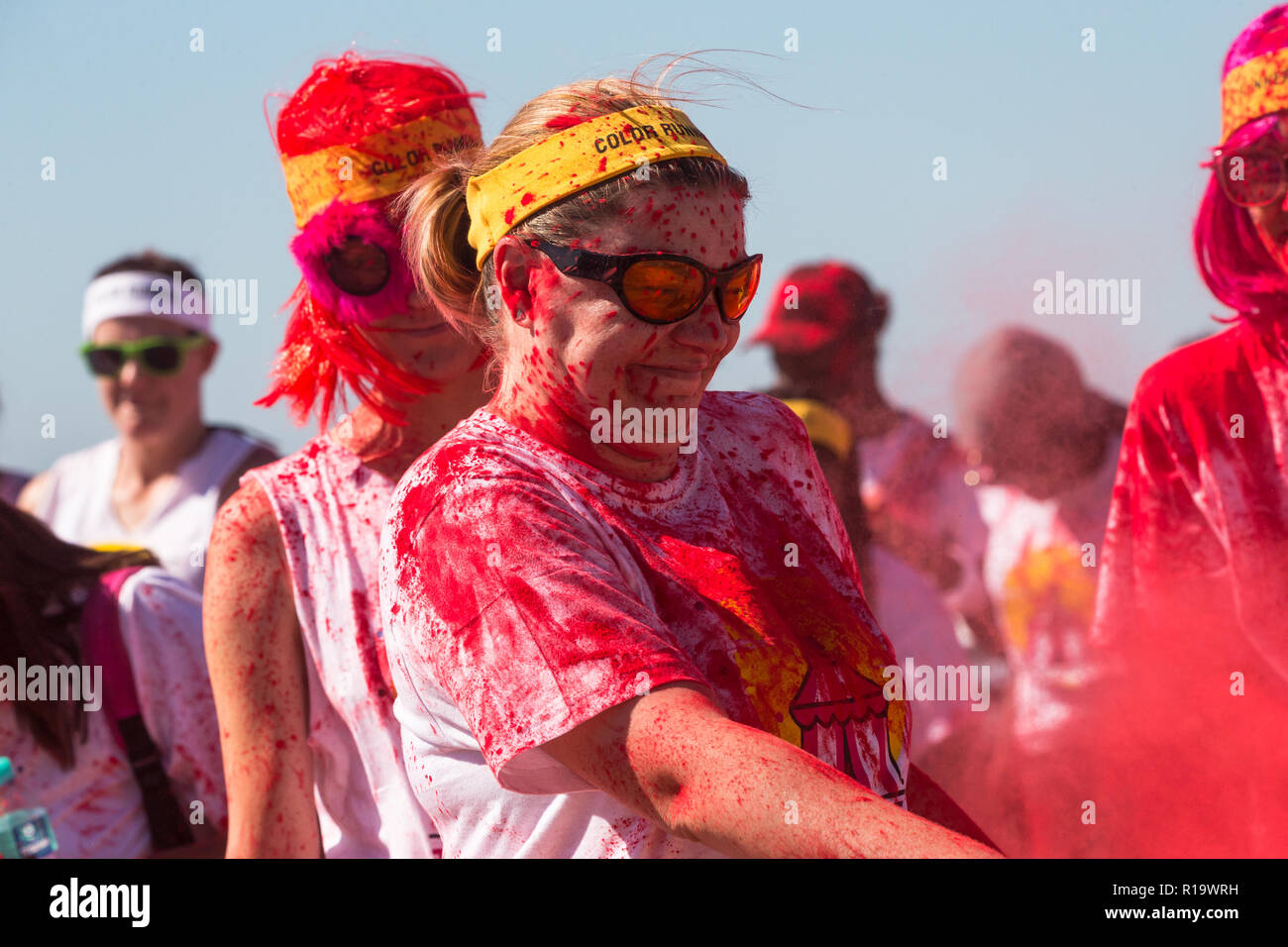 Dame de race blanche ou d'une femme portant des lunettes de soleil et un bandeau jaune recouvert de peinture en poudre rouge agitant un nuage de poudre sur quelqu'un à un événement fun run couleur d'été Banque D'Images
