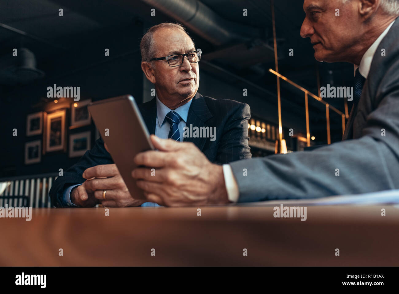 Senior businessman talking avec partenaire masculin holding digital tablet at cafe table. Deux partenaires d'affaires réunion au restaurant moderne. Banque D'Images