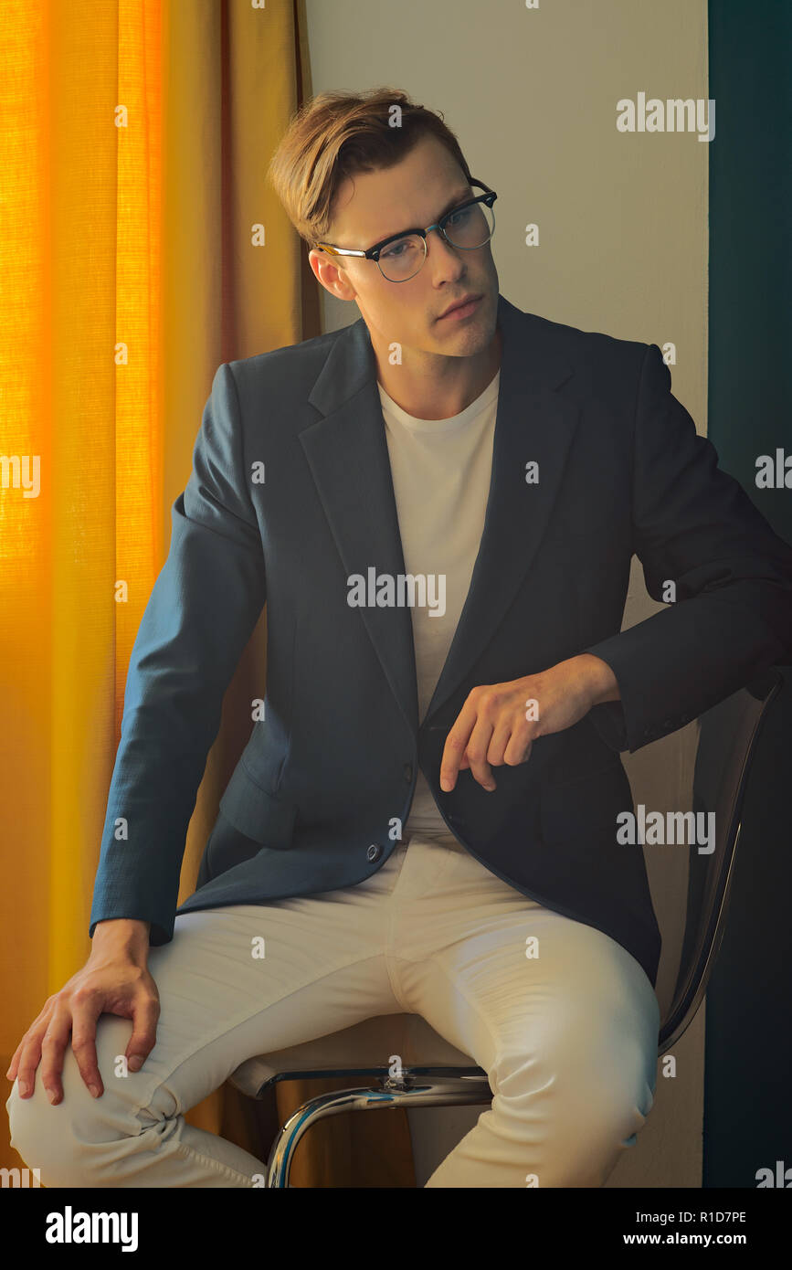 Un jeune homme, modèle masculin en costume bleu et blanc pantalon, porte une paire de lunettes vintage, assis et à l'écart, en pleine réflexion. Banque D'Images