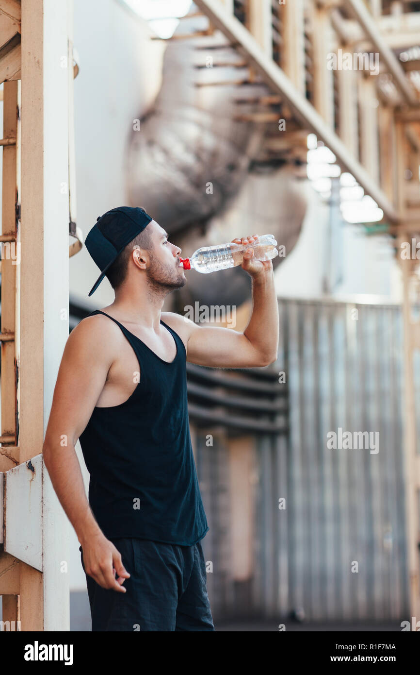 Jeune homme l'eau potable à partir de la bouteille. Sportsman se reposant après faire de l'exercice dans l'arrière-plan de la ville industrielle Banque D'Images