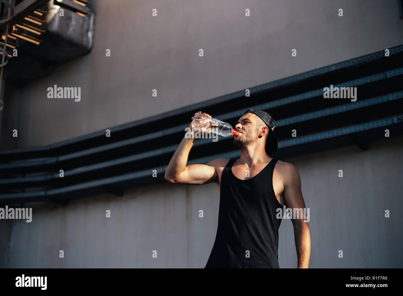 Jeune homme sportif l'eau potable à partir de la bouteille. Sportsman se reposant après faire de l'exercice dans l'arrière-plan de la ville industrielle Banque D'Images