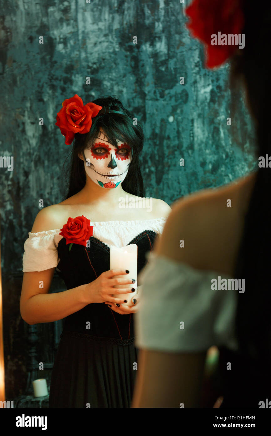 Image de sorcière femme avec un visage blanc et rouge fleur sur sa tête à côté de miroir dans la pièce sombre Banque D'Images
