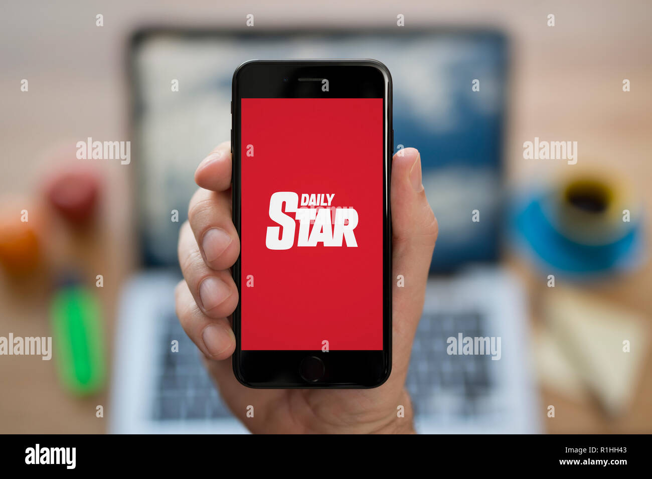 Un homme se penche sur son iPhone qui affiche le logo Daily Star, en restant assis devant son ordinateur 24 (usage éditorial uniquement). Banque D'Images