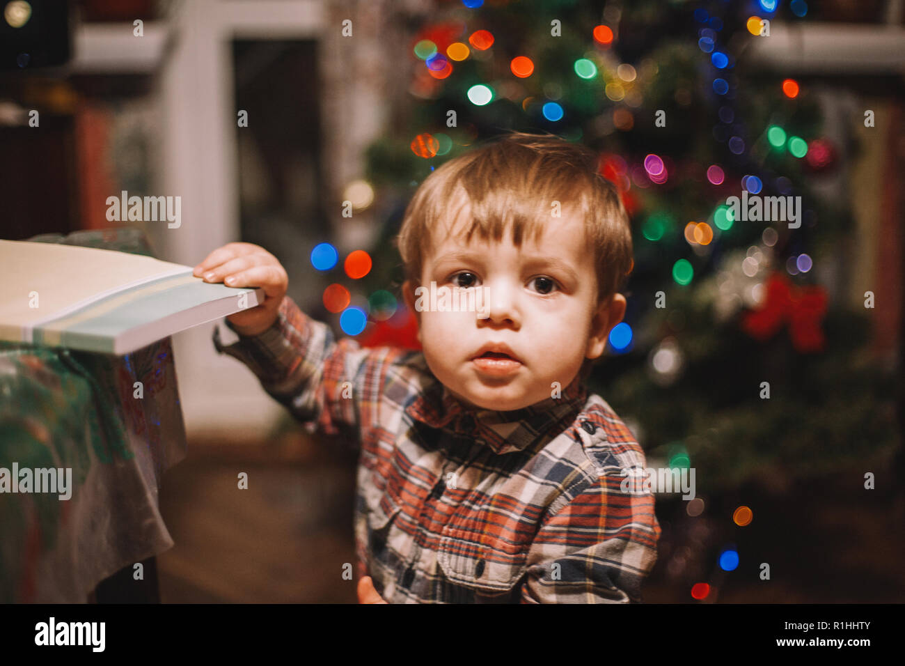 Baby Boy holding book debout contre l'arbre de Noël à la maison Banque D'Images