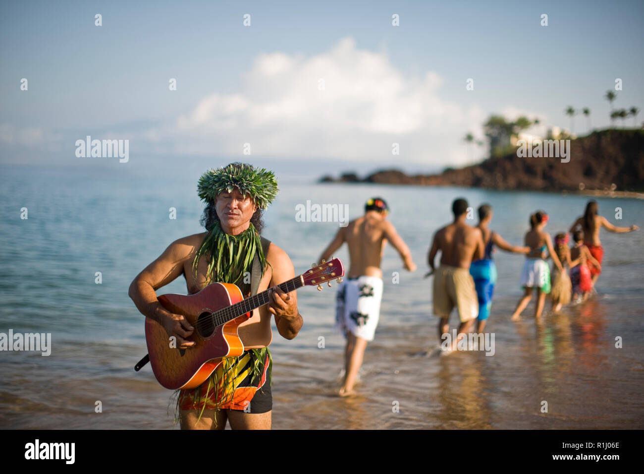 L'homme qui joue de la guitare sur la plage, avec des danseurs de hula en arrière-plan. Banque D'Images