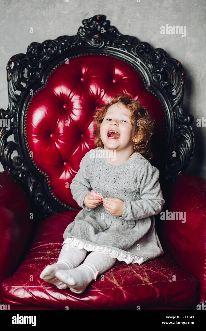 Rire fille aux cheveux bouclés en robe rouge gris assis en fauteuil. Banque D'Images