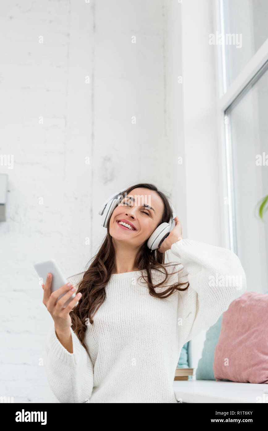 Smiling young woman listening music avec le smartphone et un casque sans fil à la maison Banque D'Images