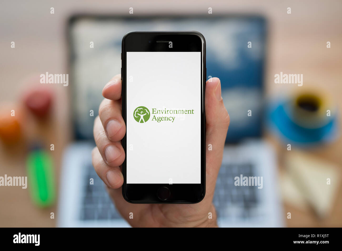Un homme se penche sur son iPhone qui affiche le logo de l'Agence de l'environnement, tout en s'assit à son bureau informatique (usage éditorial uniquement). Banque D'Images
