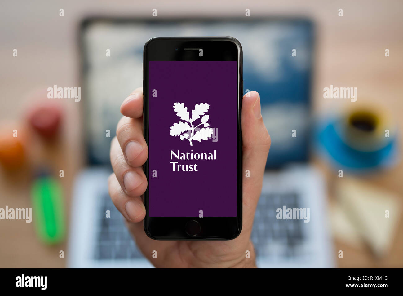 Un homme se penche sur son iPhone qui affiche le logo National Trust, tout en s'assit à son bureau informatique (usage éditorial uniquement). Banque D'Images