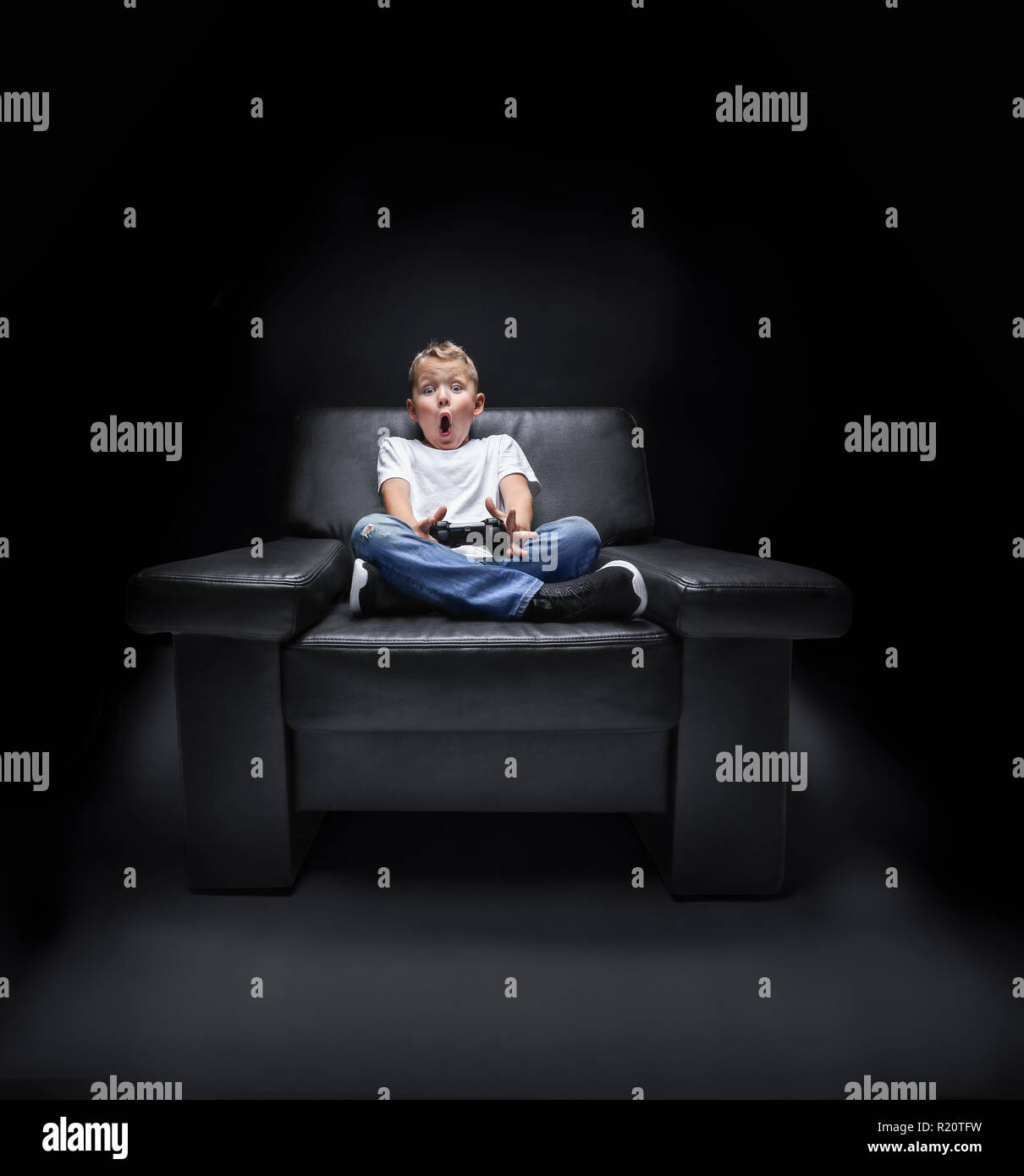 Jeune garçon en fauteuil à regarder la télévision et hurle avec le contrôleur dans sa main en face de fond noir Banque D'Images