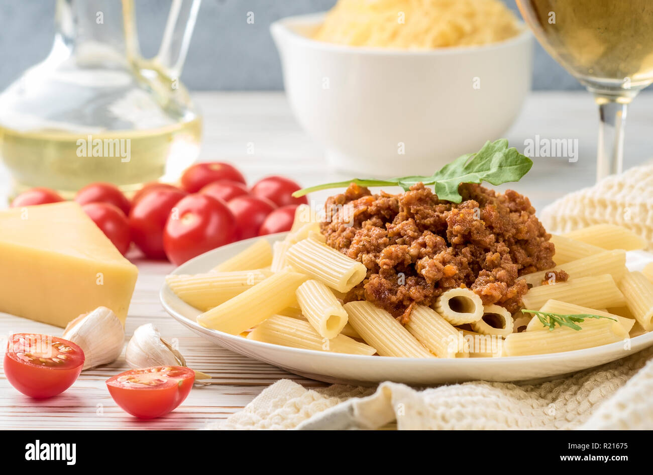 Les pâtes italiennes Penne bolognaise rigatone la viande hachée à la sauce tomate et de parmesan. Nature morte sur table en bois blanc, servi avec des tomates cerises, carafe d'huile d'olive et le verre de vin blanc Banque D'Images