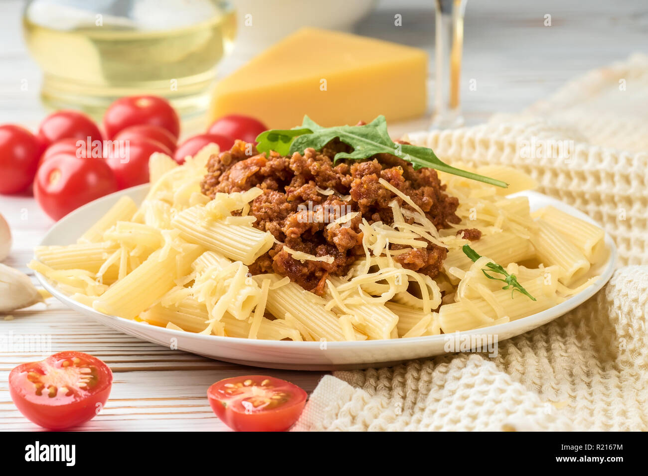 Les pâtes italiennes Penne bolognaise rigatone la viande hachée à la sauce tomate et de parmesan. Nature morte sur table en bois blanc, servi avec des tomates cerises, carafe d'huile d'olive et le verre de vin blanc Banque D'Images