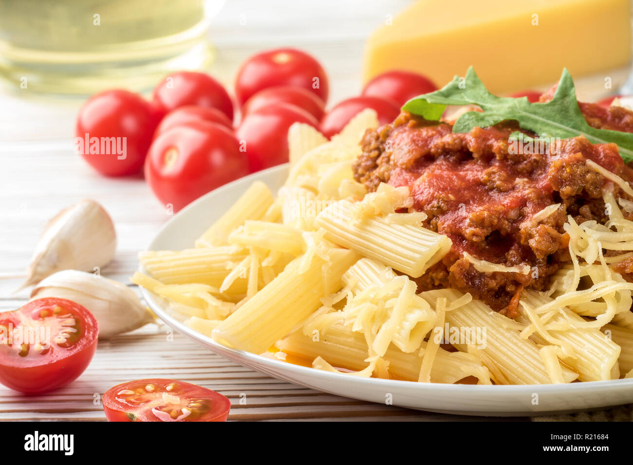 Les pâtes italiennes Penne bolognaise rigatone la viande hachée à la sauce tomate et de parmesan. Nature morte sur table en bois blanc, servi avec des tomates cerises, carafe d'huile d'olive et le verre de vin blanc Banque D'Images