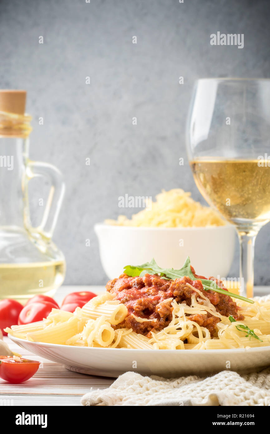 Les pâtes italiennes Penne bolognaise rigatone la viande hachée à la sauce tomate et de parmesan. Nature morte sur table en bois blanc, servi avec des tomates cerises, carafe d'huile d'olive et le verre de vin blanc Banque D'Images