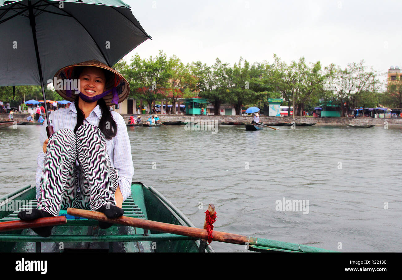 Femme sourit comme elle lignes d'un bateau avec ses pieds et est titulaire d'un parapluie sous la pluie, Tam Coc, Vietnam Banque D'Images