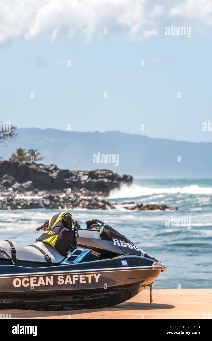 Life Guard jetski sauvetage est assis sur le sable à hawaii avec des vagues, des rochers, des maisons, et les nuages en arrière-plan. Composition verticale. Banque D'Images