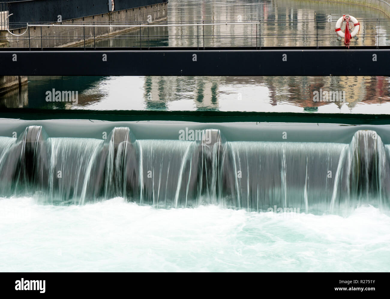 Gros plan d'une digue en béton d'origine humaine à Lucerne avec le bleu de la rivière Reuss sur elle en cascade et une passerelle métallique avec un anneau de sauvetage et d'équipement derrière Banque D'Images