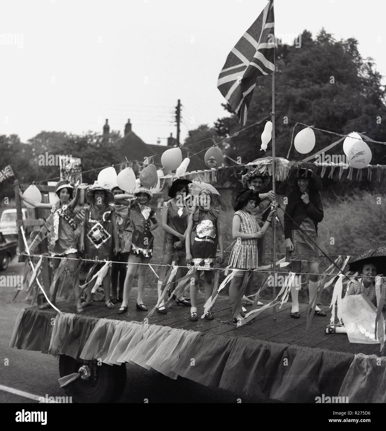 1967, jeunes filles en costumes se tenir sur l'arrière d'un char décoré dans un camion couvert de ballons et Bunting, un Union Jack flag.en tant qu'elles participent dans un village anglais carnaval de rue dans l'Oxfordshire, Angleterre, Royaume-Uni. Banque D'Images
