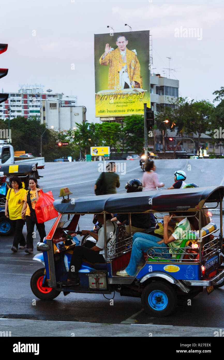Bangkok, Thaïlande : un tuk-tuk plein de passagers durs devant une avec un immense portrait du roi Bhumibol Adulyadej en centre-ville de Bangkok. Banque D'Images
