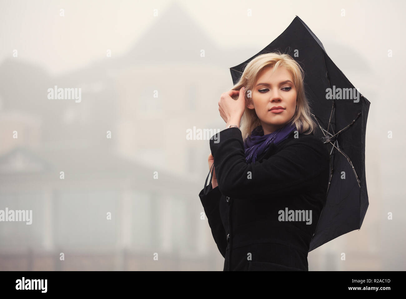Jeune femme blonde avec parapluie marche dans un brouillard. Modèle de mode élégant manteau noir classique en piscine Banque D'Images