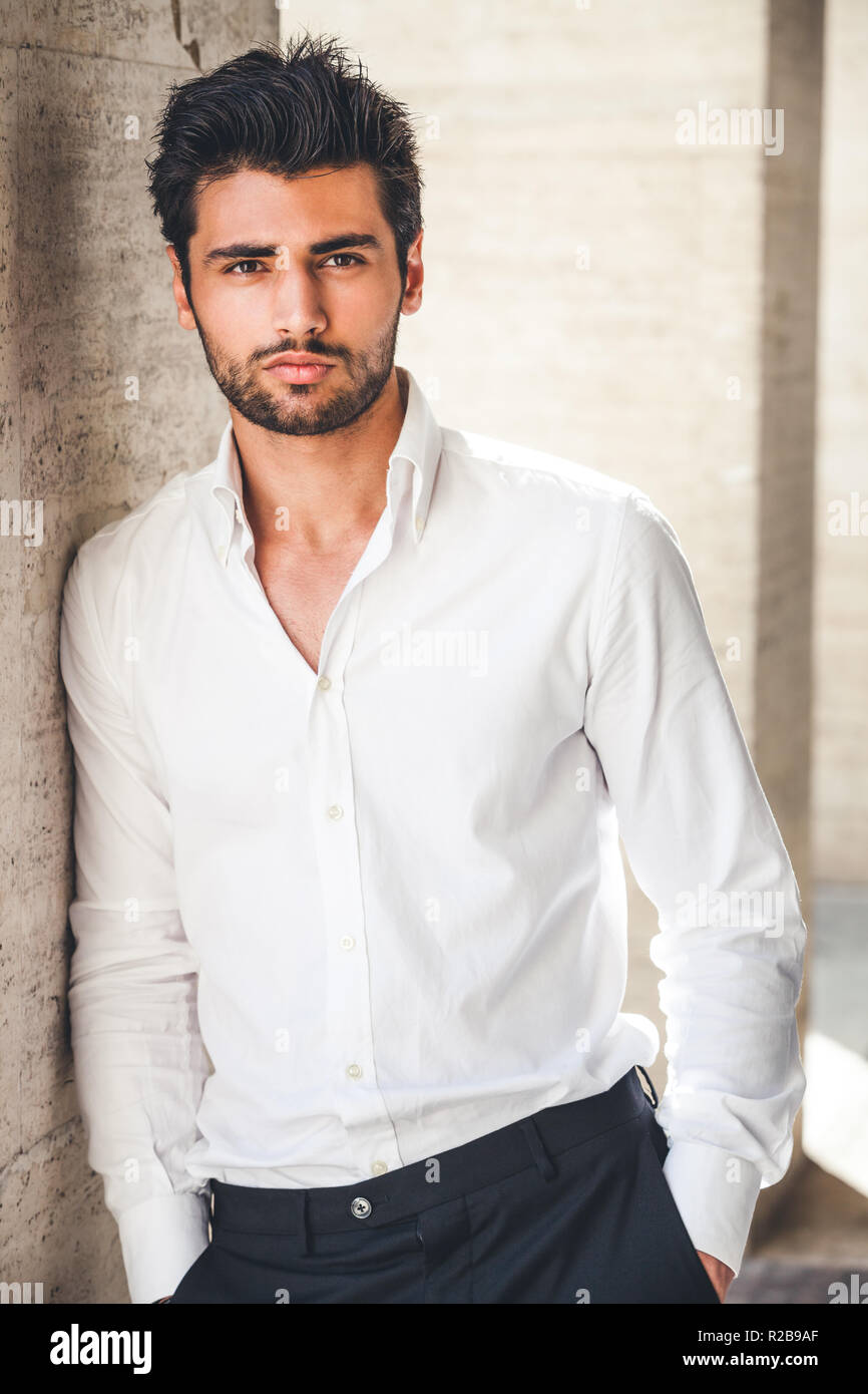 Portrait de jeune homme séduisant en chemise blanche à l'extérieur. Belle apparence avec des cheveux et la barbe. Se penchant d'un côté sur un mur. Banque D'Images