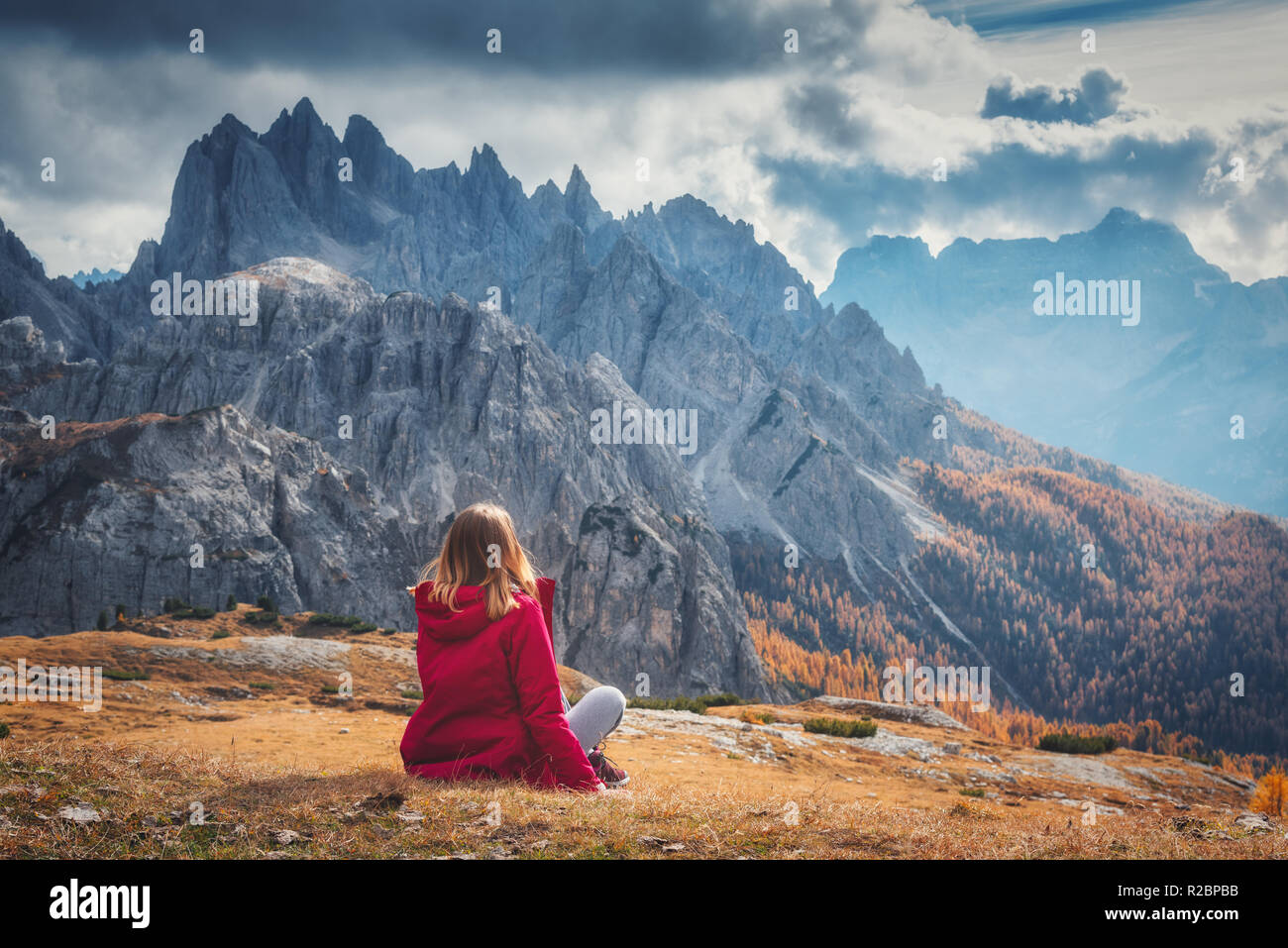 Jeune femme est assise sur la colline contre les montagnes majestueuses au coucher du soleil en automne en Dolomites, Italie. Paysage avec fille, ciel nuageux, orange gra Banque D'Images