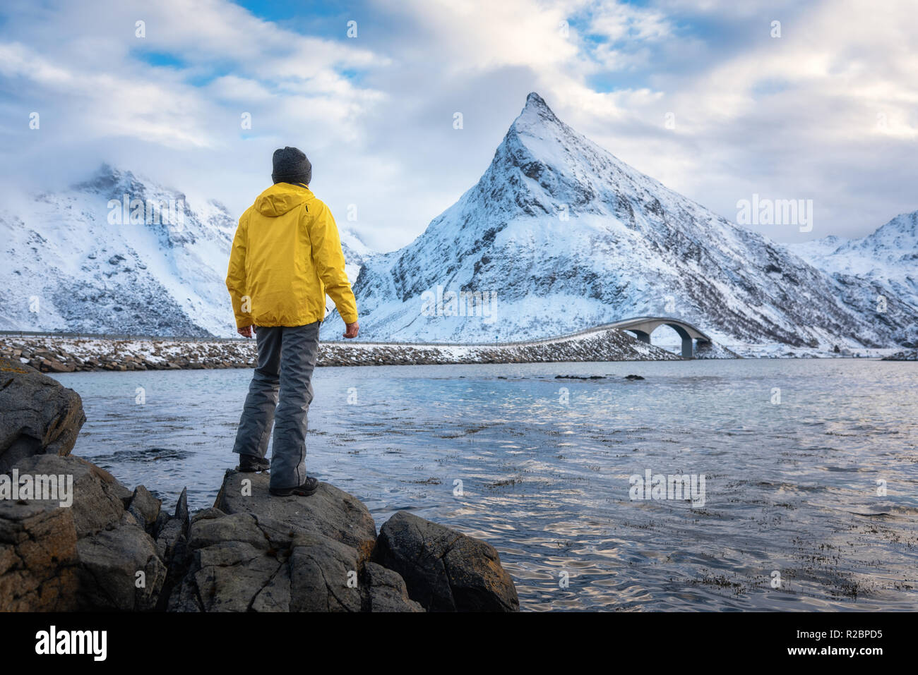 L'homme sportif dans la région de Yellow Jacket debout sur la pierre sur le littoral contre les montagnes de neige et ciel nuageux au coucher du soleil en hiver. Paysage avec traveler, wat Banque D'Images