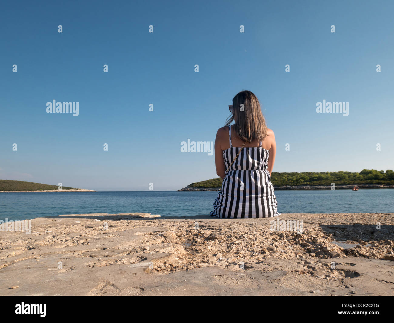 Belle femme en noir et robe blanche assise seule sur un quai en pierre près de la mer sur la chaude journée d'été Banque D'Images