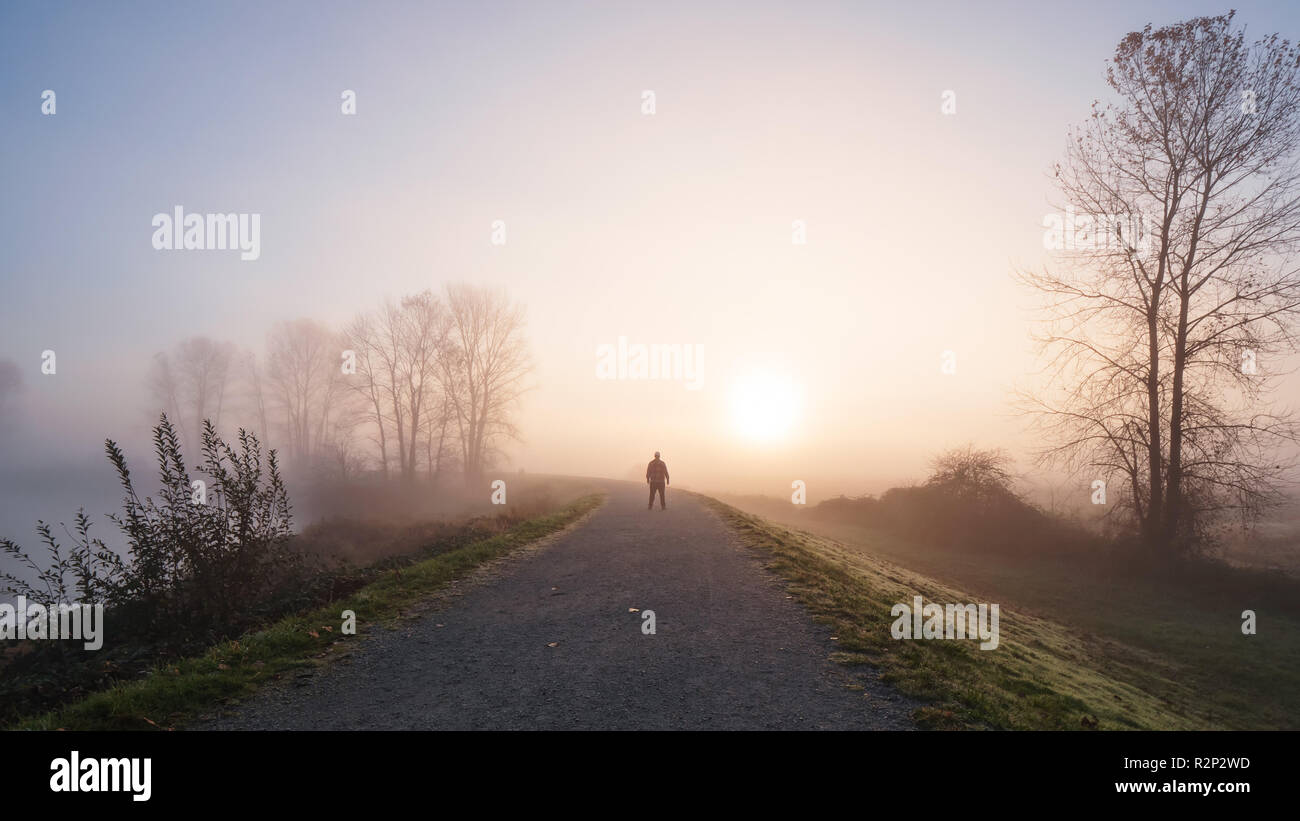 Homme debout sur un long chemin de gravier avec un lever de soleil brumeux au loin. Banque D'Images Homme debout sur un long chemin de gravier avec un lever de soleil brumeux au loin. Banque D'Images
