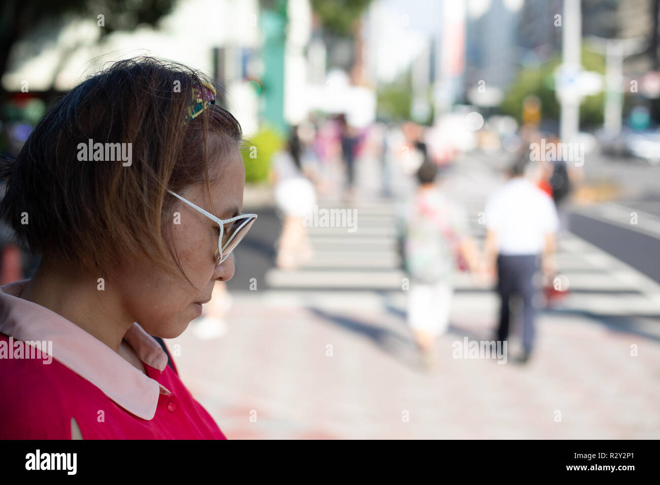 Portrait of mature femme chinoise de Taiwan seul se tenant dans l'ombre à la recherche vers le bas tandis que les gens traversent une grande ville intersection en plein soleil Banque D'Images