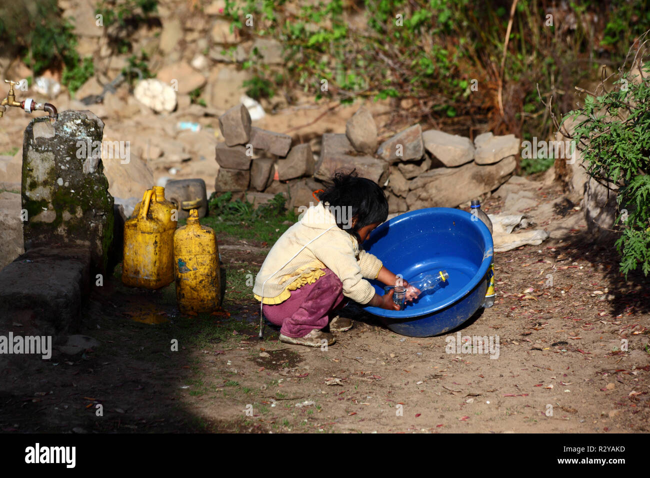 Jeune enfant autochtone jouant avec une bouteille en plastique, un bol en plastique bleu et de l'eau dans un village rural andin, Bolivie Banque D'Images