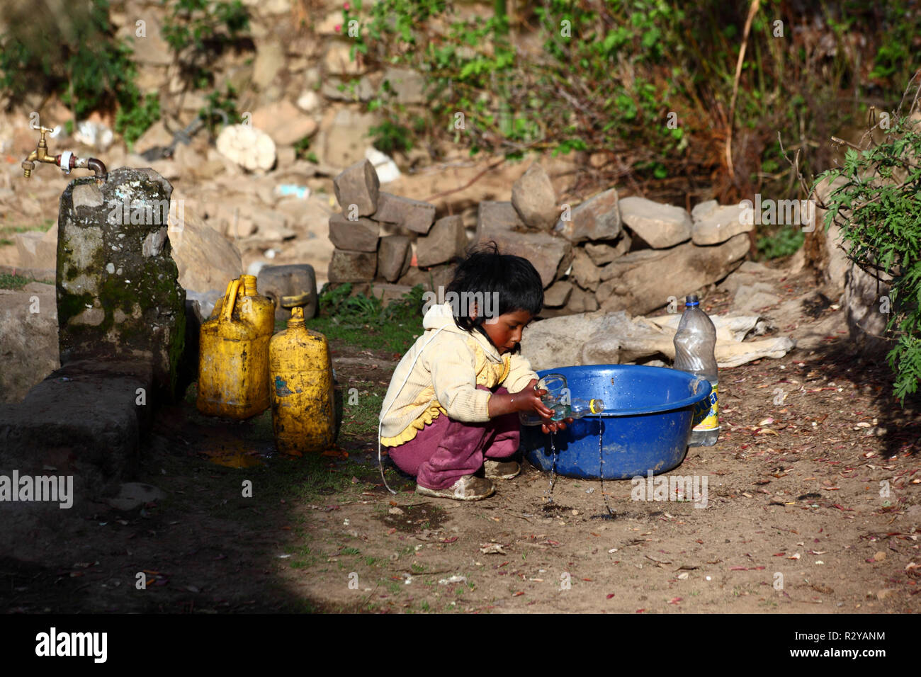 Jeune enfant autochtone jouant avec une bouteille en plastique, un bol en plastique bleu et de l'eau dans un village rural andin, Bolivie Banque D'Images