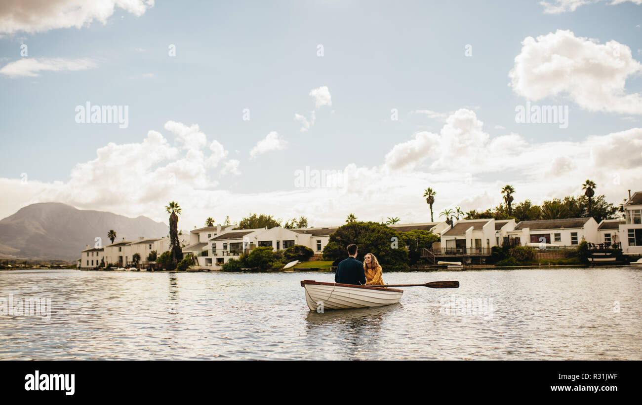 Vue panoramique d'un couple romantique à bord d'un lac. Jeune couple rowing leur bateau dans un lac avec des maisons en arrière-plan. Banque D'Images