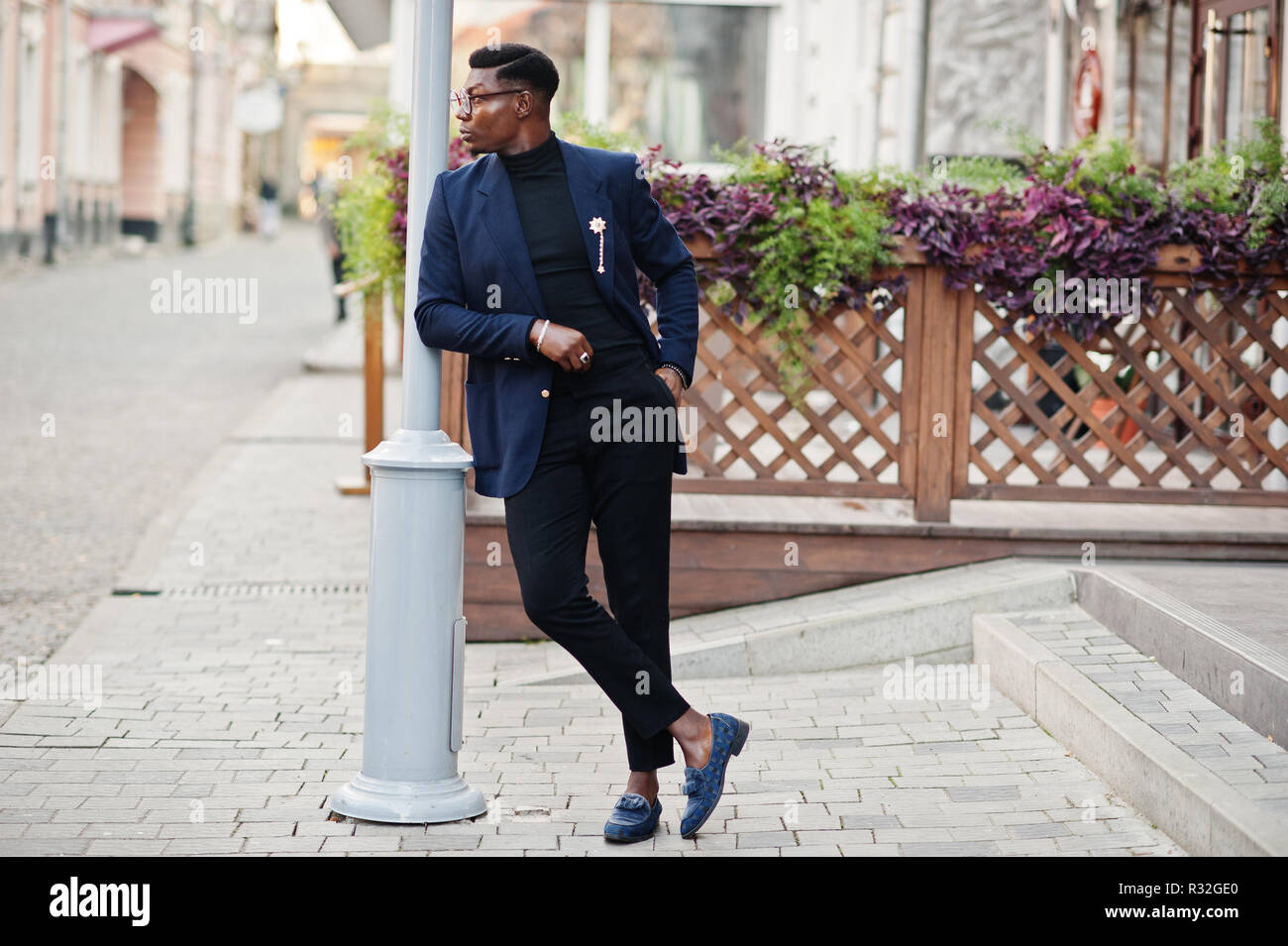 un homme afro-américain à la recherche étonnante porte un blazer bleu avec  une broche, un col roulé noir et des lunettes posées dans la rue. mec noir  à la mode avec téléphone