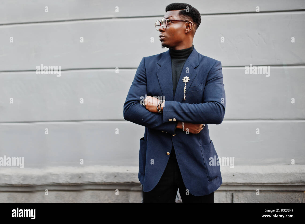 un homme afro-américain à la recherche étonnante porte un blazer bleu avec  une broche, un col roulé noir et des lunettes posées dans la rue. mec noir  à la mode avec téléphone