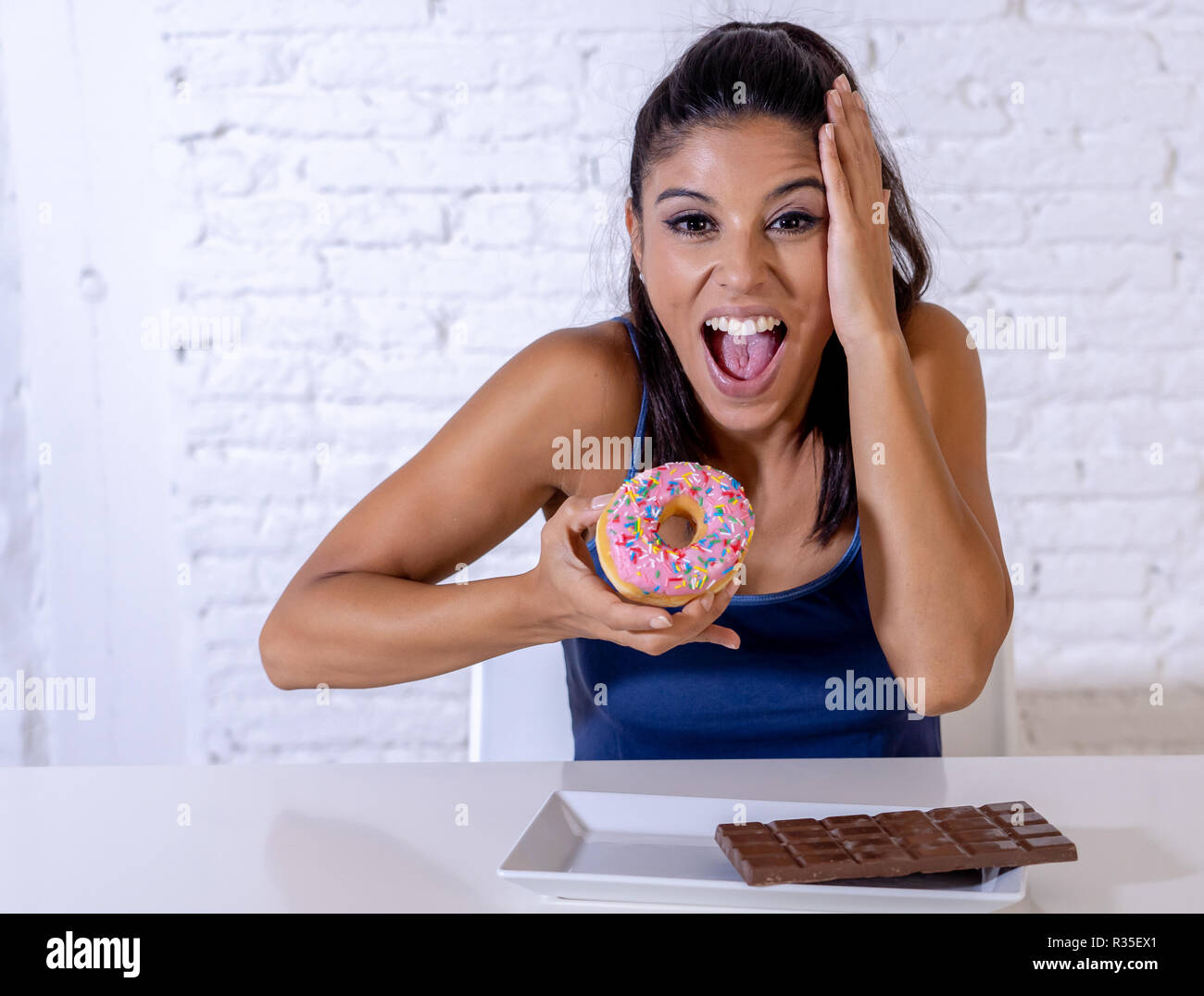 Young attractive woman sitting at table latine sur le point de manger du chocolat et des beignets à la excité et heureux en plus de l'alimentation, le sucre et l'unhealt chunky Banque D'Images