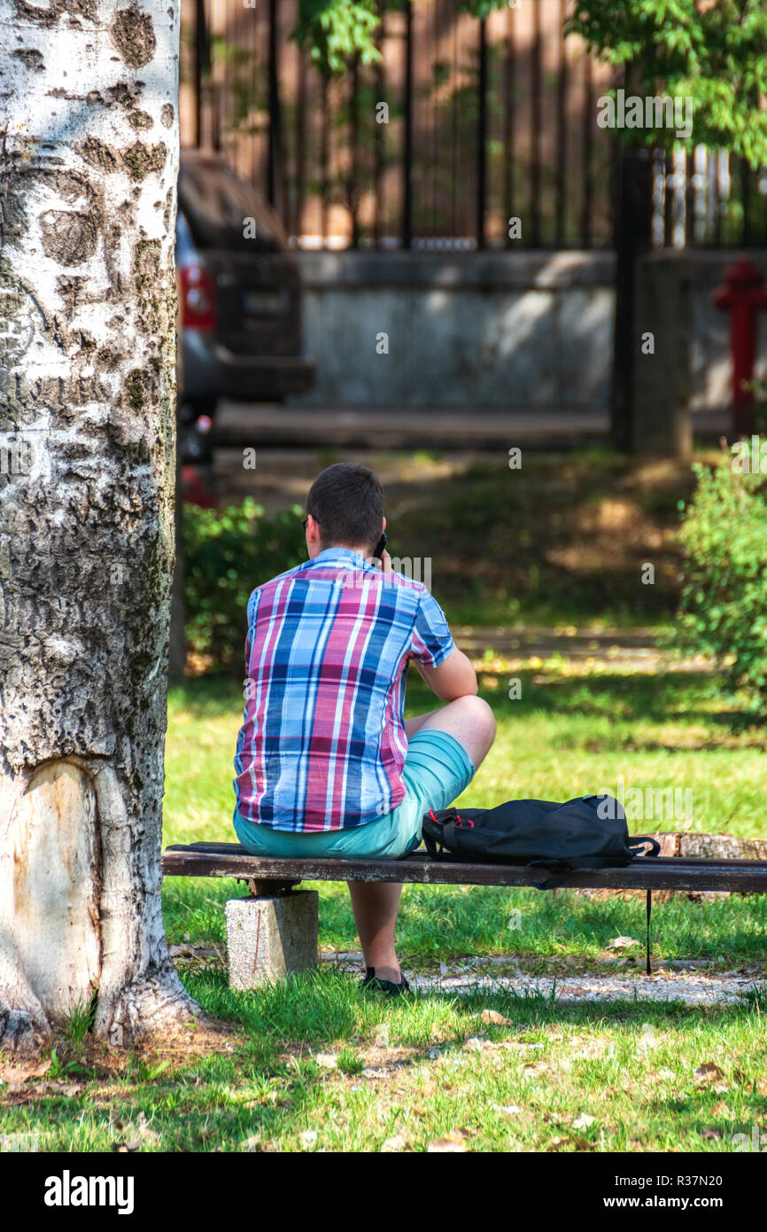 Vue depuis l'arrière sur un jeune homme assis sur un banc, à parler au téléphone dans un parc de la ville dans la ville de Szeged, Hongrie. Banque D'Images