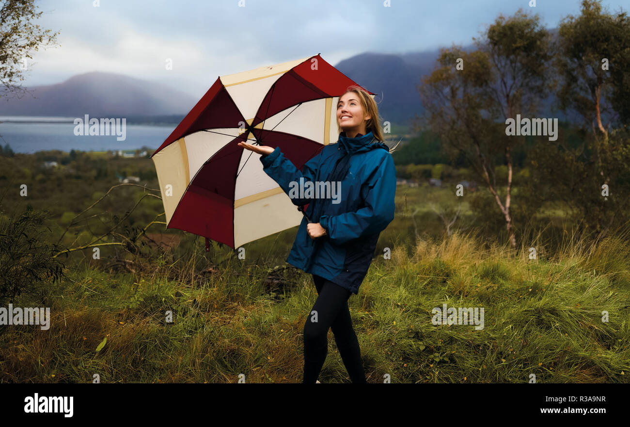 Jolie femme aux cheveux blonds, debout avec un parapluie en plein air et jouit de la pluie. Le port de big blue Jacket. Un paysage extraordinaire sur l'arrière-plan Banque D'Images