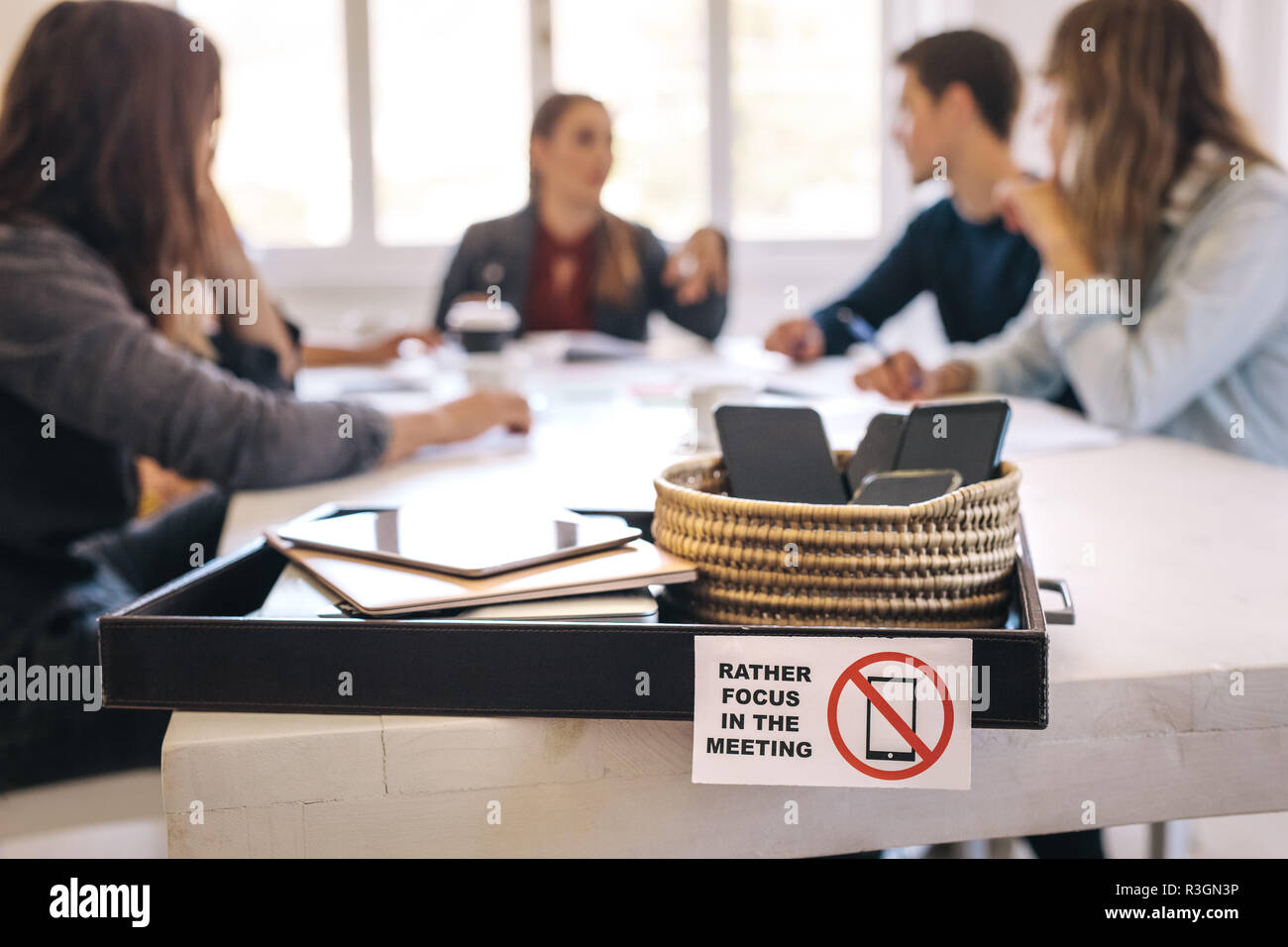 Les téléphones portables, ordinateurs portables et tablettes numériques dans un bac sur table de réunion avec des gens assis à l'arrière et discuter de travail. Réunion sans techno Banque D'Images