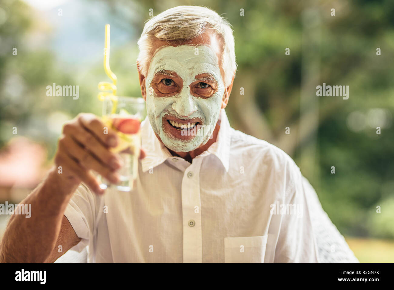 Funny man avec de l'argile masque du visage tenant un verre de jus. Traitement facial spa homme ayant pris sa retraite à l'âge. Banque D'Images