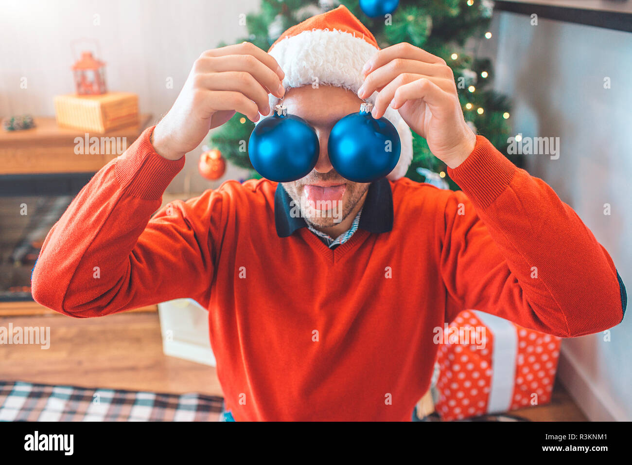 Jeune homme drôle détient sur les jouets les yeux avec les mains. Ils sont bleu. Il joue avec elle. Guy porte pull de Noël et un chapeau. Il est heureux Banque D'Images