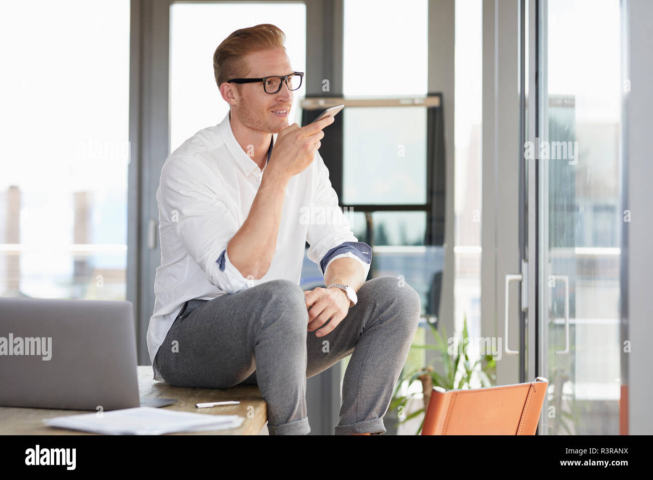 Smiling businessman sitting on desk in office using smartphone Banque D'Images