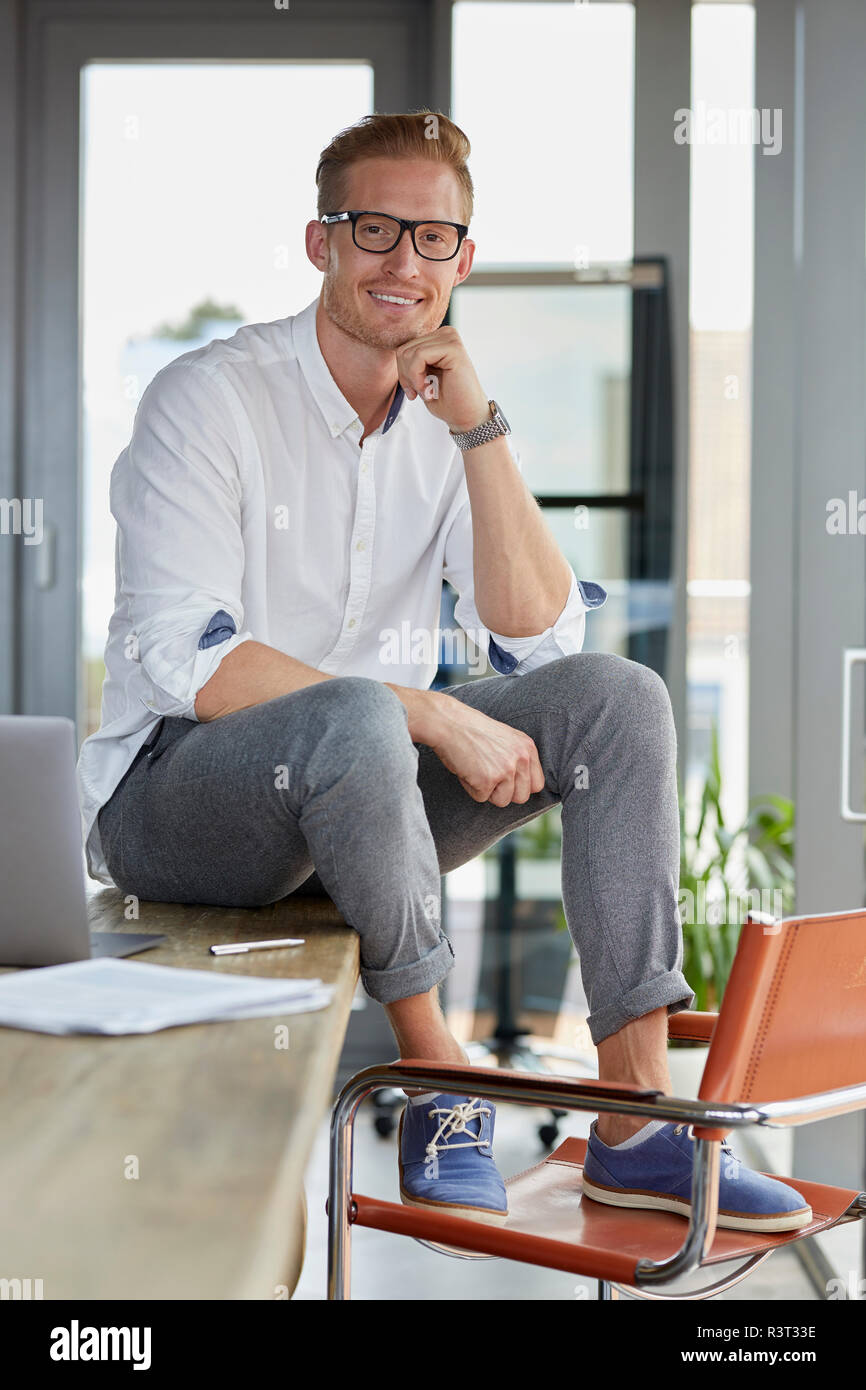 Portrait of smiling businessman with laptop sitting on desk in office Banque D'Images