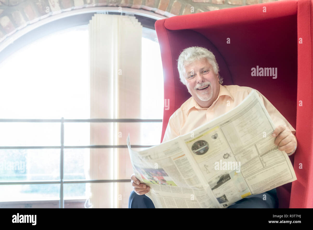 Senior lachender Vergnügt mit einem roten Sessel Zeitung. Grevenbroich, Nordrhein-Westfalen, Deutschland Banque D'Images