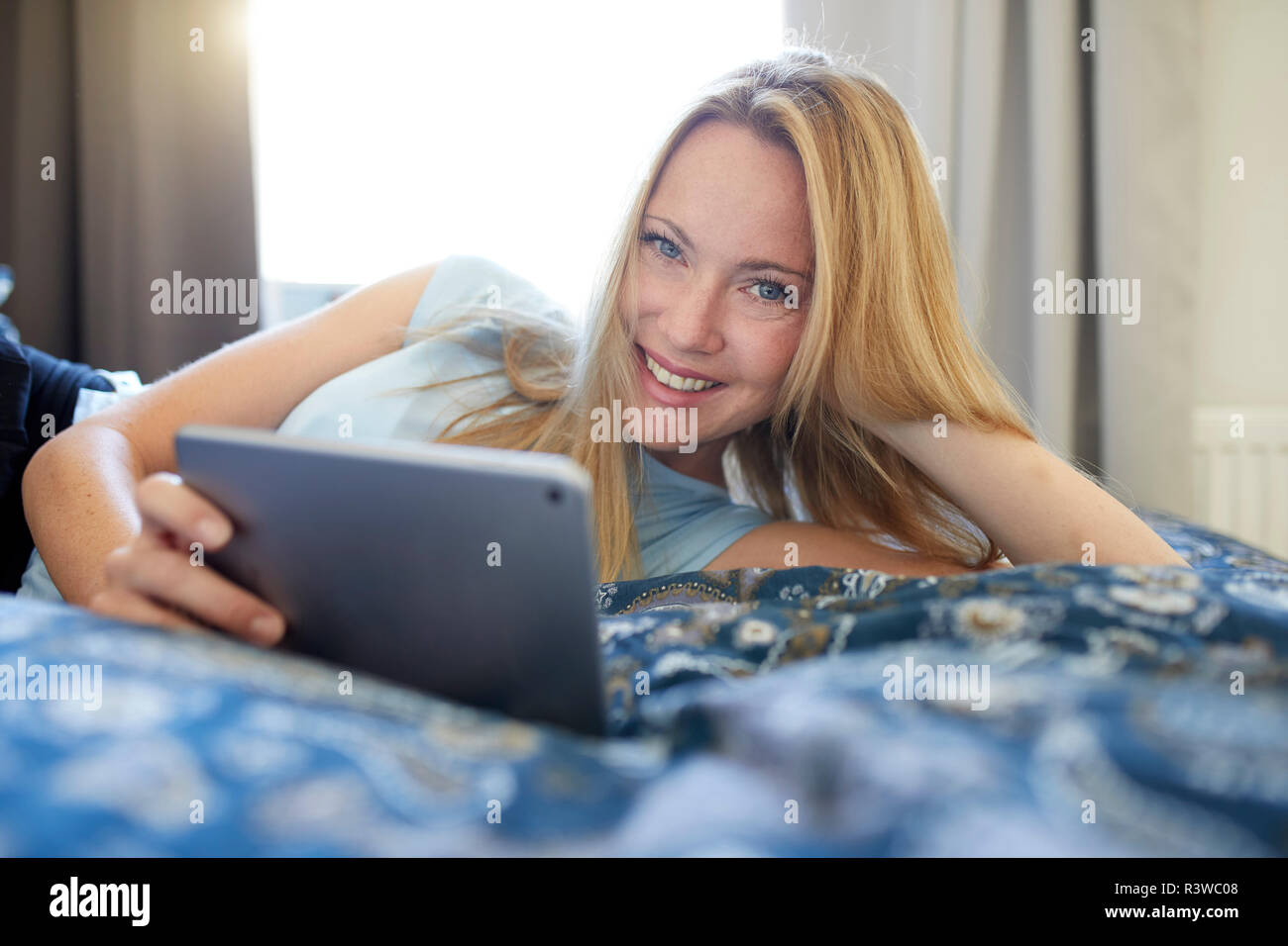 Laughing young woman lying on bed at home, using digital tablet Banque D'Images