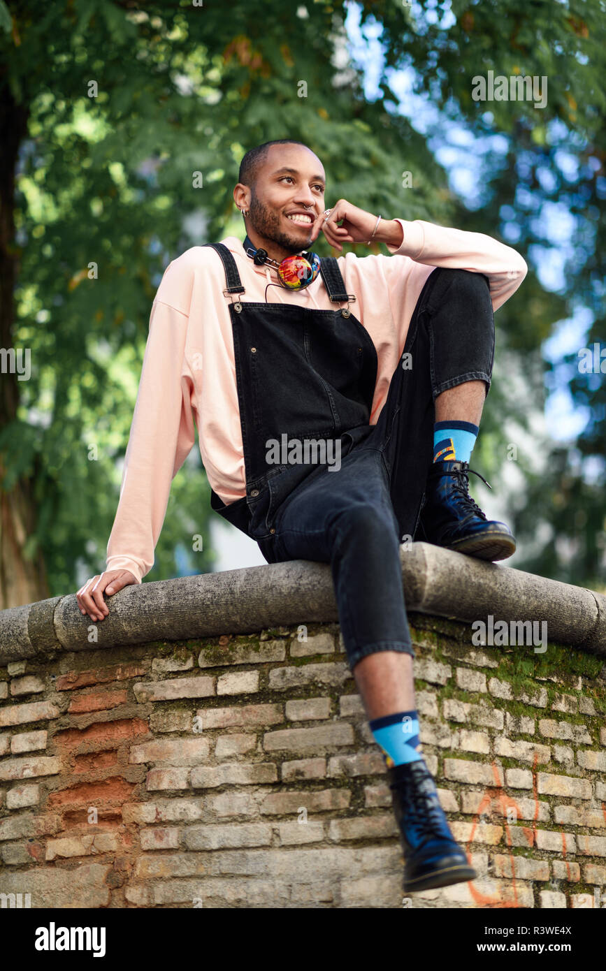 Jeune homme noir portant des vêtements décontractés et des écouteurs assis sur fond urbain. Happy African guy avec les salopettes en plein air Banque D'Images