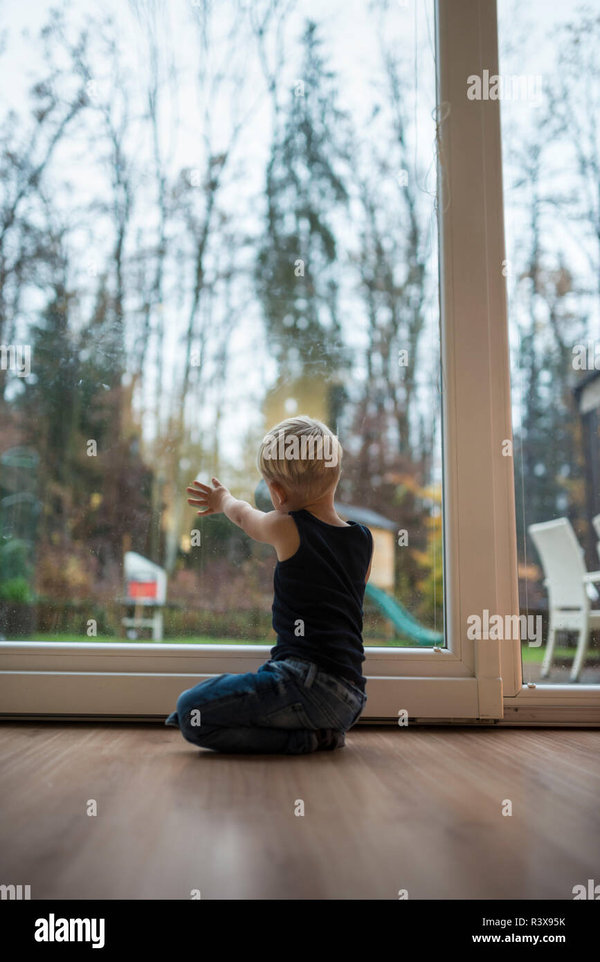 Petit garçon blond à genoux sur un sol en bois à la maison en face d'une porte patio en verre donnant sur le jardin avec sa main sur la vitre en lumière du soir. Banque D'Images