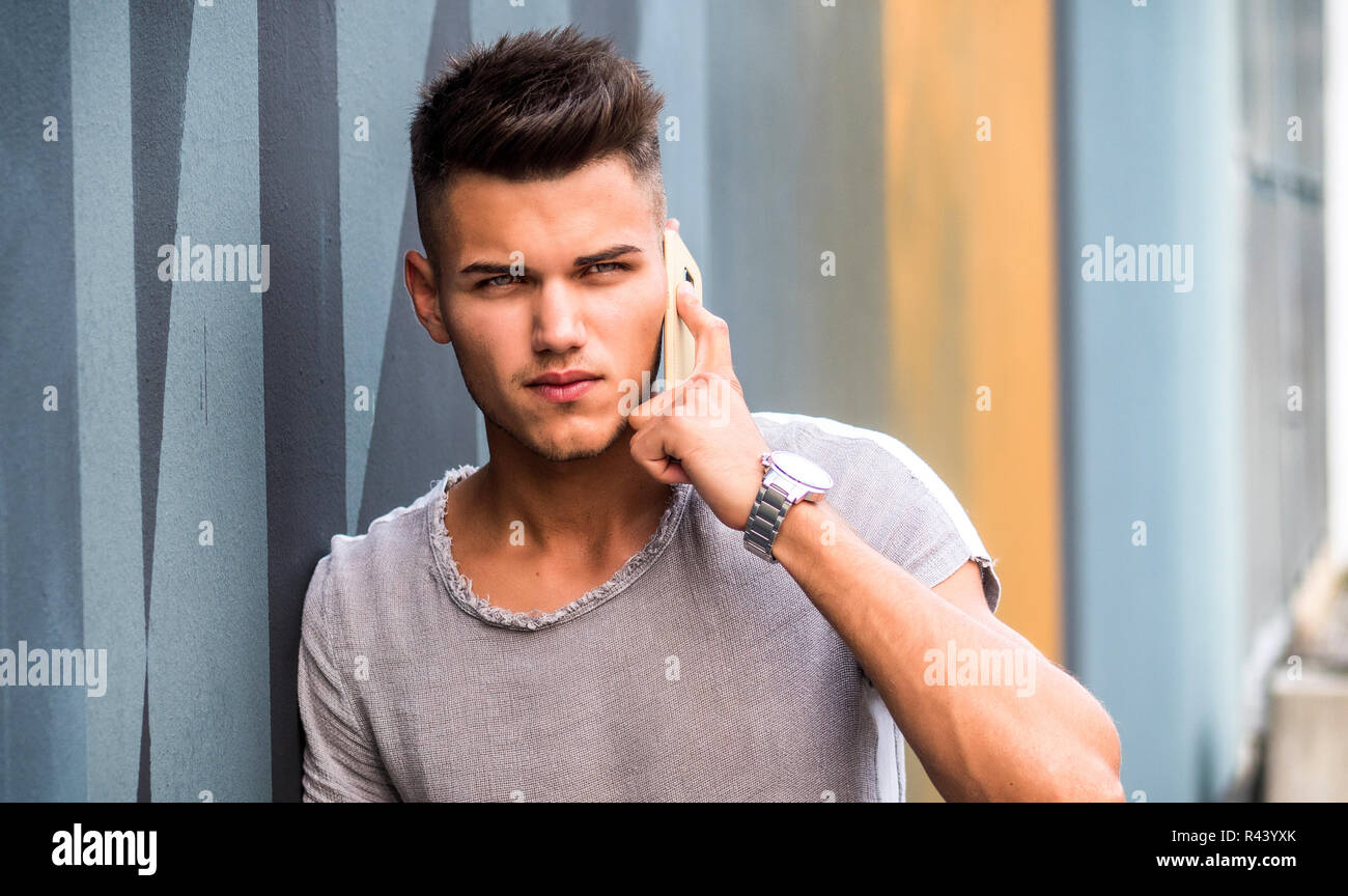 Beau jeune homme debout à l'extérieur dans l'environnement urbain à côté de mur coloré, talking on cell phone Banque D'Images