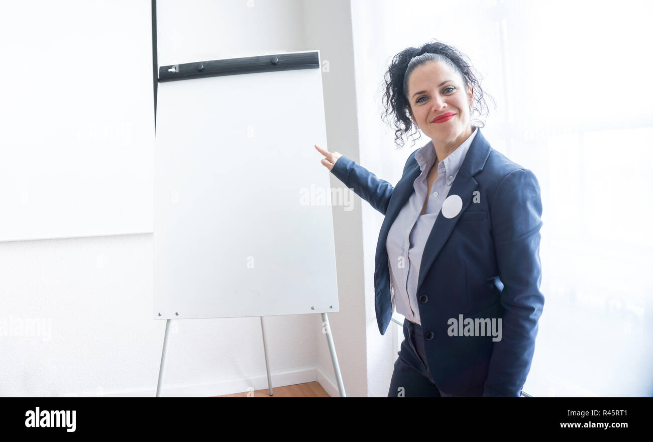 Portrait d'une femme normale d'âge moyen aux yeux bleus, la peau blanche et les cheveux noirs habillés de façon professionnelle dans un bureau regardant la caméra pointant sur un tableau blanc vide. Souriant avec le rouge à lèvres rouge Banque D'Images